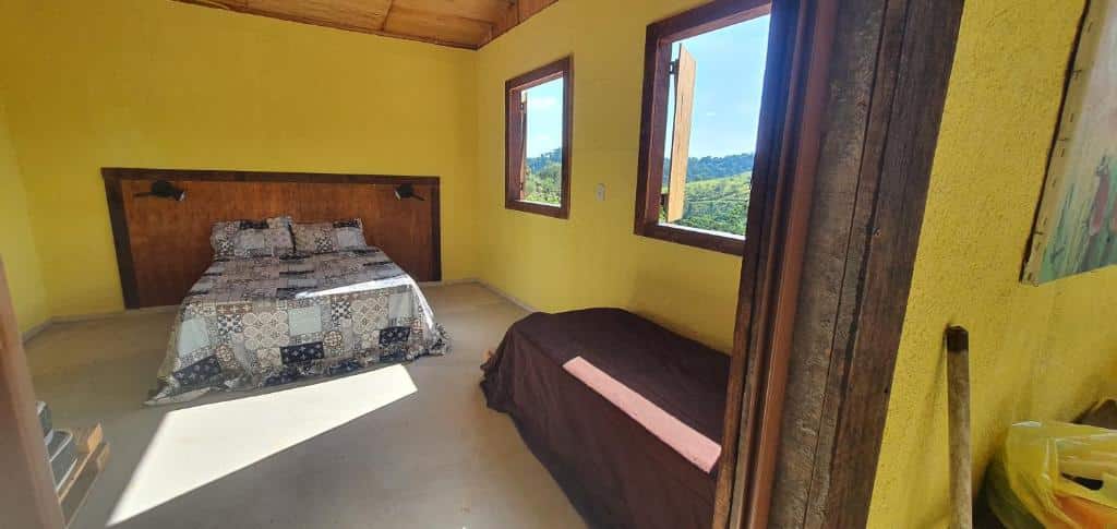 Um quarto simples no Recanto das Acácias com uma cama de casal, uma cama de solteiro, paredes amarelas e duas janelas abertas com vista para a vegetação externa.