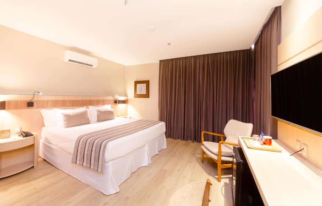 Quarto moderno no Ritz Copacabana Boutique Hotel com uma cama bem arrumada, TV, poltrona, escrivaninha e cortinas escuras do chão ao teto, para representar hotéis em Copacabana.