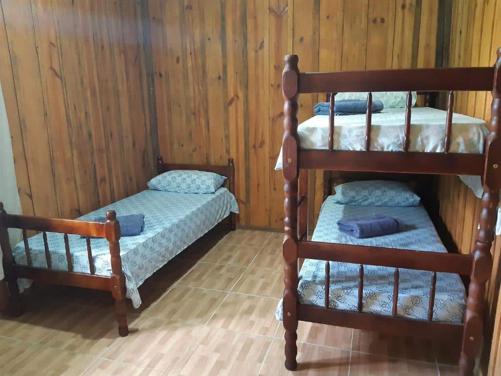 Um quarto na Pousada Serra Verde em Morretes com paredes de madeira, uma cama de solteiro e um beliche de madeira, todos com roupas de cama estampadas e toalhas dobradas, para representar pousadas perto de Curitiba.