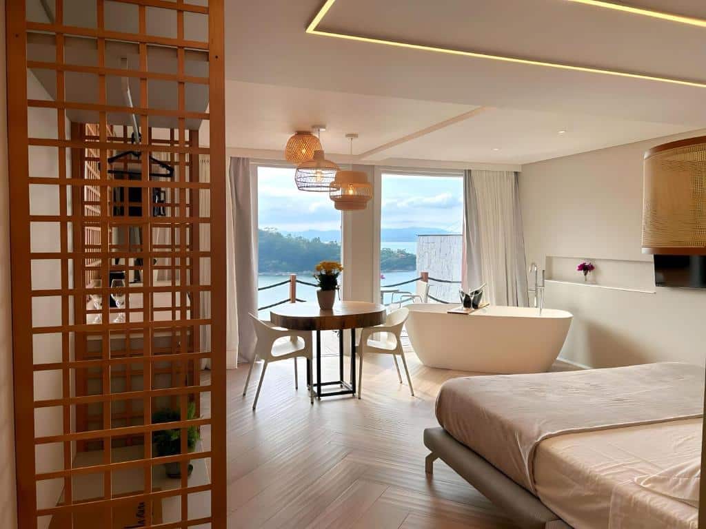 Quarto no Studio Vista do Mar com uma cama, banheira independente, mesa de jantar, cadeiras, janela grande e vista panorâmica para o exterior, para representar pousadas em Governador Celso Ramos.