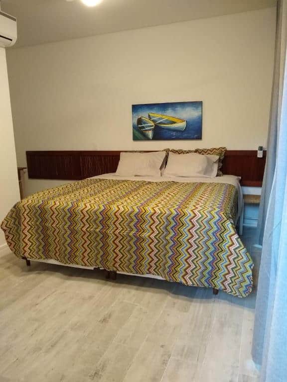 Uma cama bem arrumada nas Suítes KM18praia com roupas de cama com padrão em ziguezague em um quarto com piso de madeira clara e uma pintura de barcos acima da cabeceira, para representar pousadas na Barra do Una.