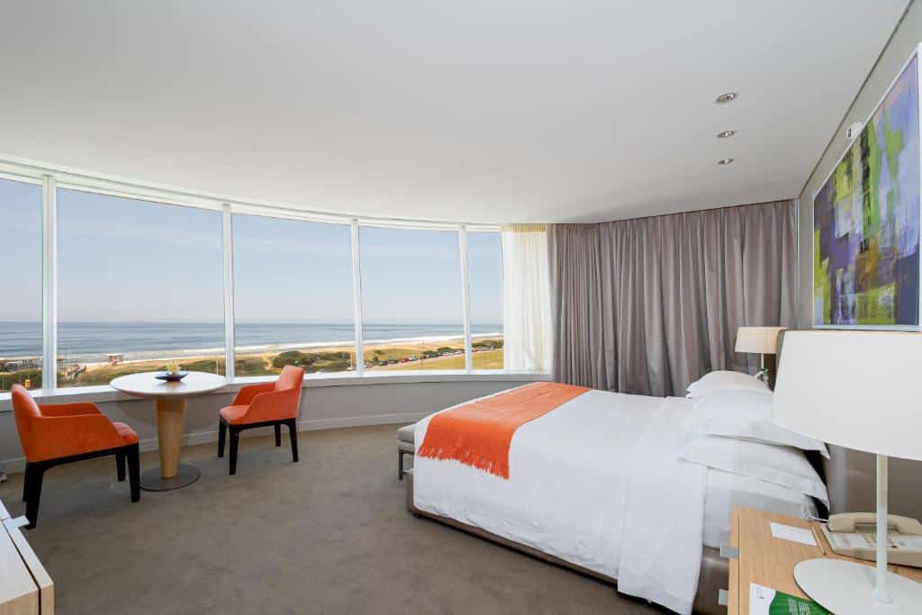Quarto no The Grand Hotel com uma cama de casal, duas cadeiras laranjas em uma mesa redonda, janelas grandes, vista para o mar e decoração moderna, para representar onde ficar em Punta Del Este.