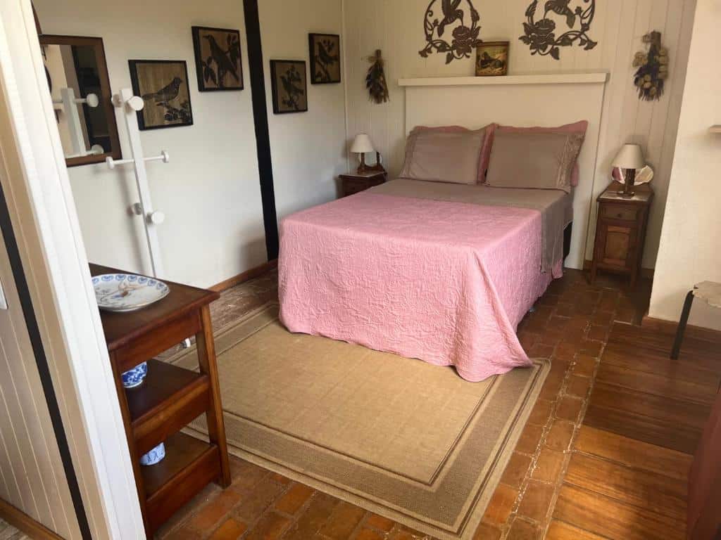 Uma cama bem arrumada com uma capa rosa em um quarto pequeno com móveis de madeira, arte na parede e um tapete bege no chão, no The Lodge em Teresópolis, para representar chalés românticos no Rio de Janeiro.