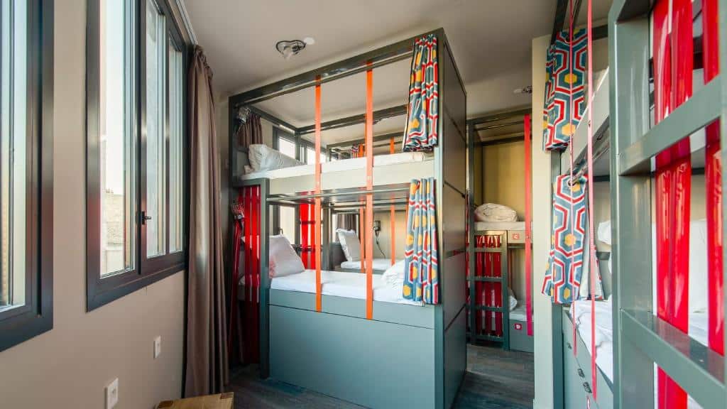 Um pequeno dormitório com dois conjuntos de beliches, cortinas coloridas e janelas grandes que permitem a entrada de luz natural no Les Piaules - Belleville.