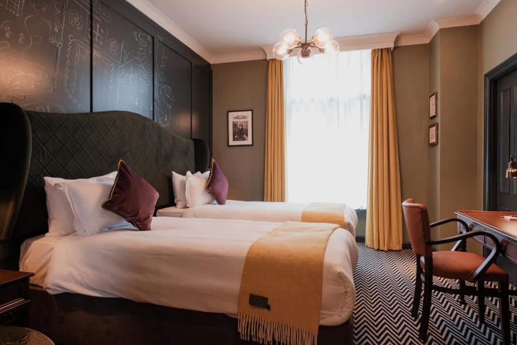 Um quarto no The Sanctuary House Hotel com duas camas de solteiro, cobertores e travesseiros amarelos, uma escrivaninha, cadeira, janela com cortinas amarelas e arte na parede, para representar hotéis perto da London Eye.