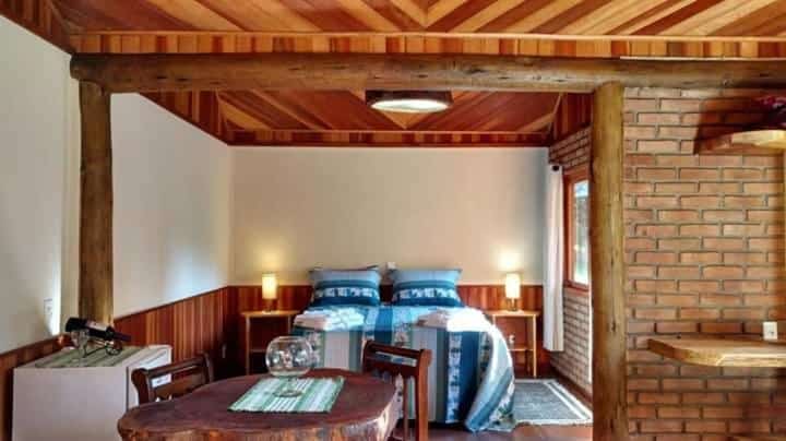 Um quarto rústico com teto de madeira, parede de tijolos, cama de casal e uma pequena mesa de jantar com duas cadeiras no Chalé Truta da Floresta em Visconde de Mauá.