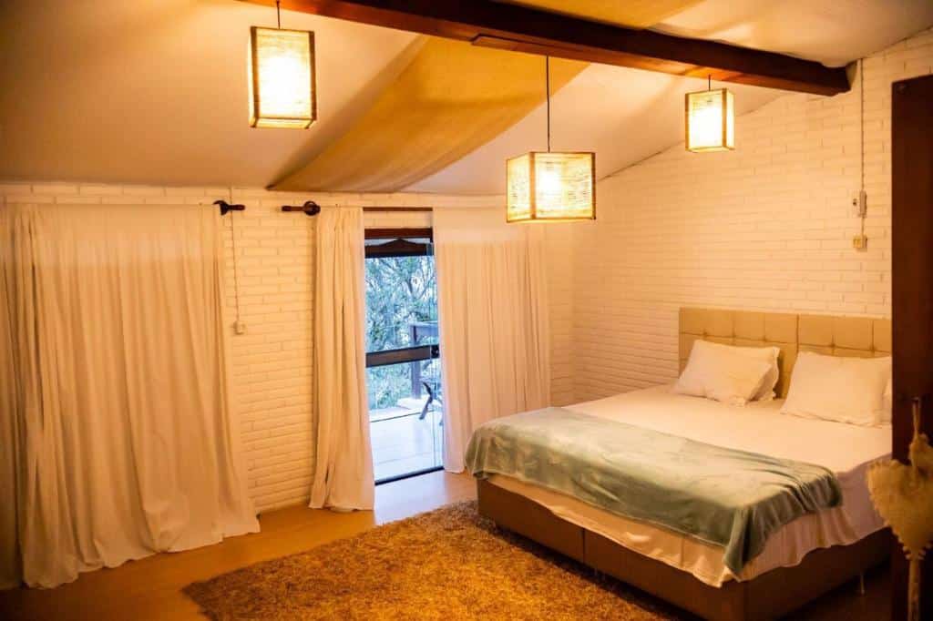 Quarto na Villa dos Ganchos Hospedaria com cama de casal, paredes de tijolos brancos, tapete bege, luzes penduradas e janela grande com cortinas, para representar pousadas em Governador Celso Ramos.