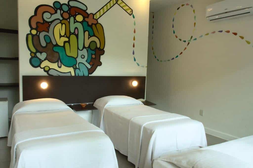 Três camas de solteiro com lençóis brancos em um quarto com arte colorida na parede e uma unidade de ar-condicionado split, na Villa dos Graffitis Pousada, para representar pousadas em Morro de São Paulo.