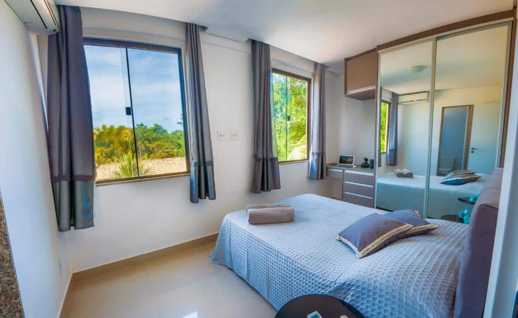 Quarto no Village Imbassaí bem iluminado com duas janelas, cortinas cinza, uma cama com roupa de cama cinza, um espelho e azulejos de cor clara no piso, para representar airbnb em Imbassaí.