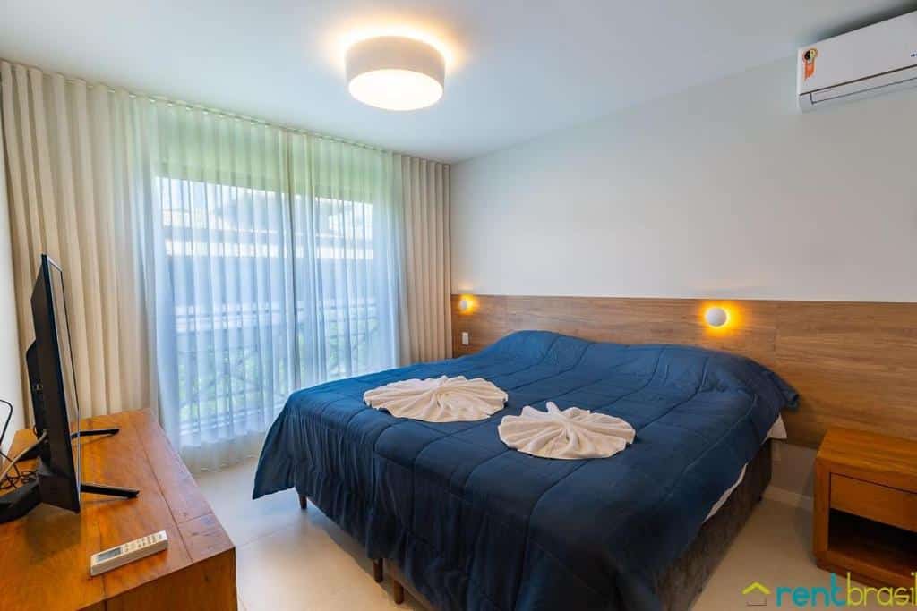 Um quarto no Ykutiba Imbassaí - Apartamento 3 Quartos Superior - I104 com uma cama de casal, roupa de cama azul, luzes de cabeceira, uma TV em uma mesa de madeira e uma grande janela com cortinas, para representar airbnb em Imbassaí.