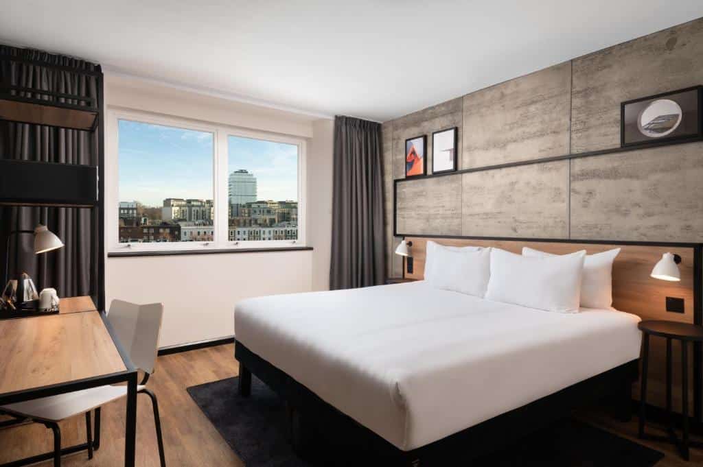 Quarto no ibis London Earls Court com uma cama de casal, escrivaninha, luminárias, arte na parede e uma janela com vista para os prédios da cidade, para representar hotéis ibis em Londres