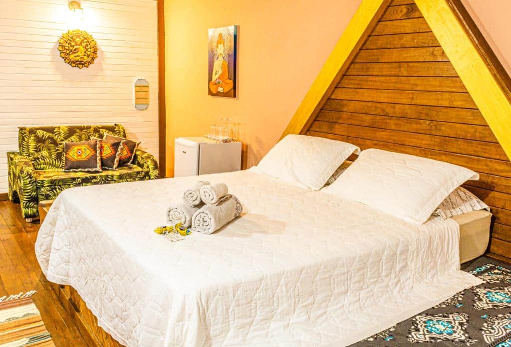Um quarto aconchegante na Pousada Casa do Oscar com uma cama branca, toalhas enroladas, um pequeno frigobar, um sofá estampado e paredes de madeira em formato de A, para representar pousadas em Govenador Celso Ramos.