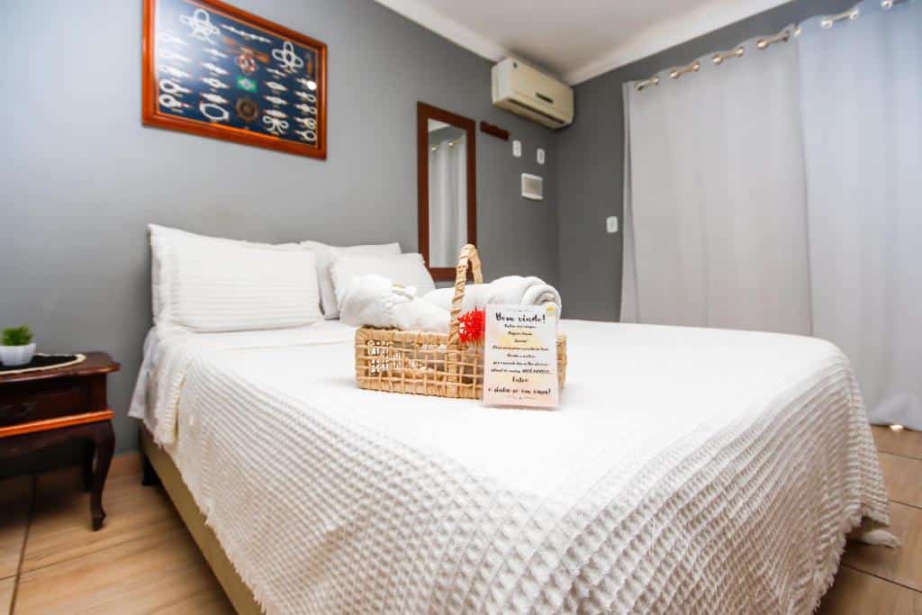Uma cama bem arrumada na Pousada Maresia Costa Azul com lençóis brancos, uma cesta de toalhas e um bilhete de boas-vindas em um quarto moderno e bem iluminado.