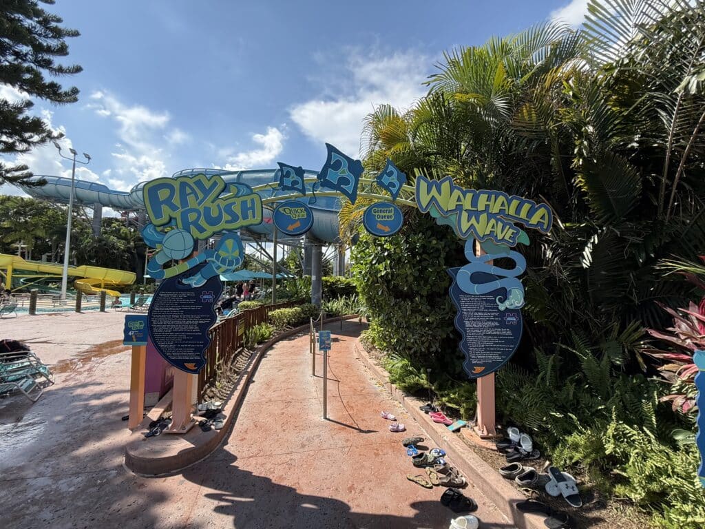 Placas de entrada para os passeios aquáticos Ray Rush e Walhalla Wave com sapatos alinhados no chão e plantas tropicais nas proximidades.
