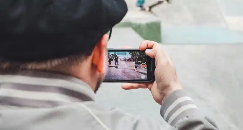 Uma pessoa de boné filma um skatista em um skatepark usando um smartphone.