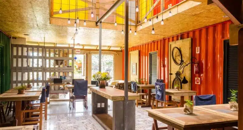 Café com mesas de madeira, luzes penduradas, plantas e uma bicicleta montada em uma parede laranja em um espaço moderno e bem iluminado, no Alma Brava, para representar pousadas em Itajaí.