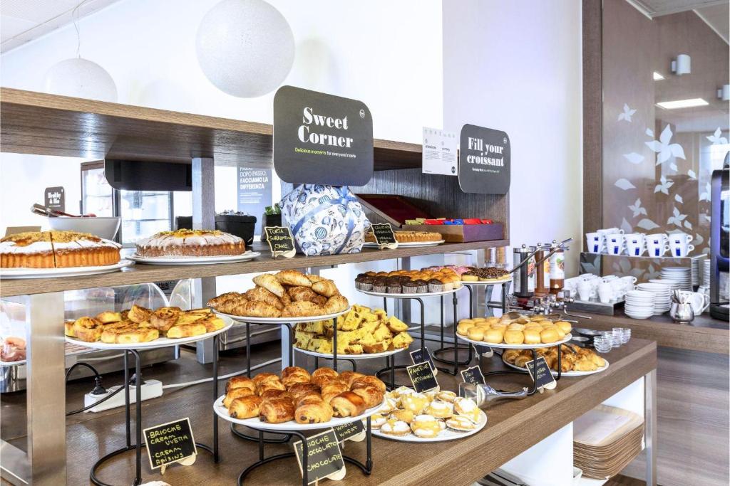 Um bufê de café-da-manhã self-service com doces, bolos e croissants dispostos em bandejas em camadas sob placas identificadas, na propriedade do B&B Hotel Milano Portello.
