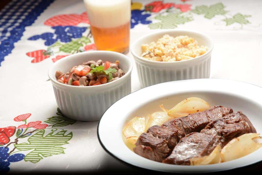 Um prato com bife grelhado e cebolas, uma tigela de ovos mexidos, uma tigela de feijão com legumes e um copo de cerveja, servido no Mangue Seco Suítes & Bistrô no Mangue Seco.