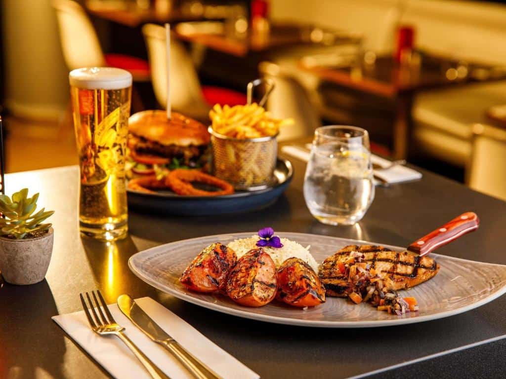 Um prato de frango grelhado e legumes, um hambúrguer com batatas fritas, um copo de cerveja e água em uma mesa de restaurante, servido no ibis London Blackfriars.