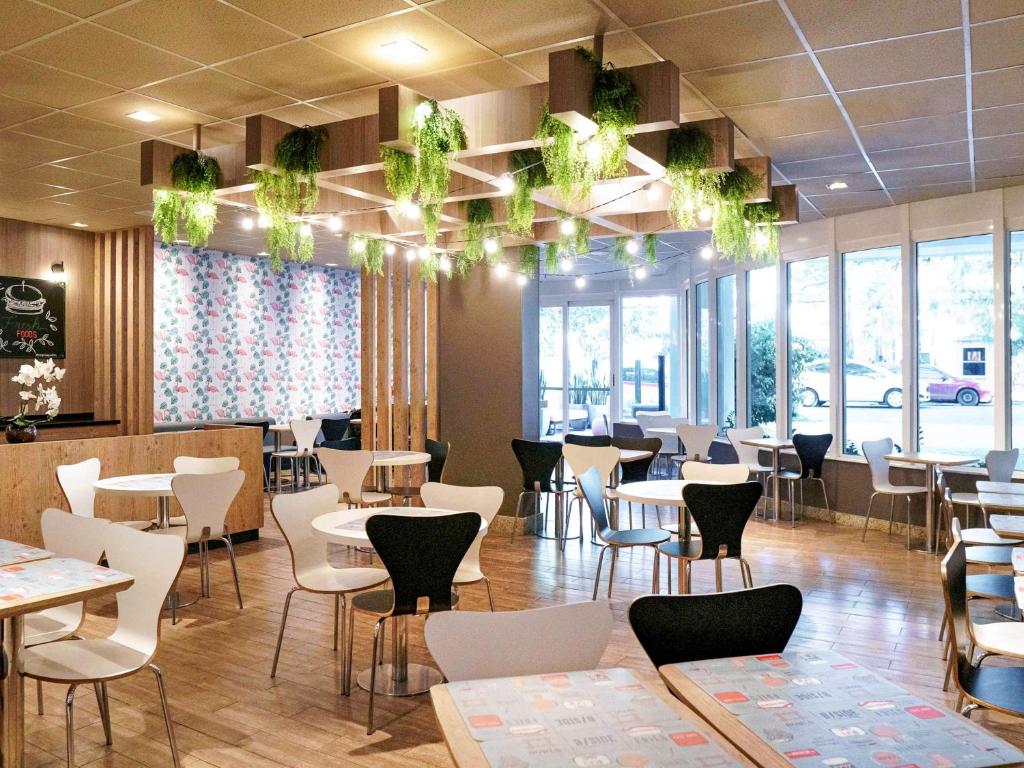 Café moderno com piso de madeira, cadeiras brancas e pretas, mesas redondas e luzes no teto com plantas verdes penduradas, na propriedade do ibis Porto Alegre Moinhos de Vento.