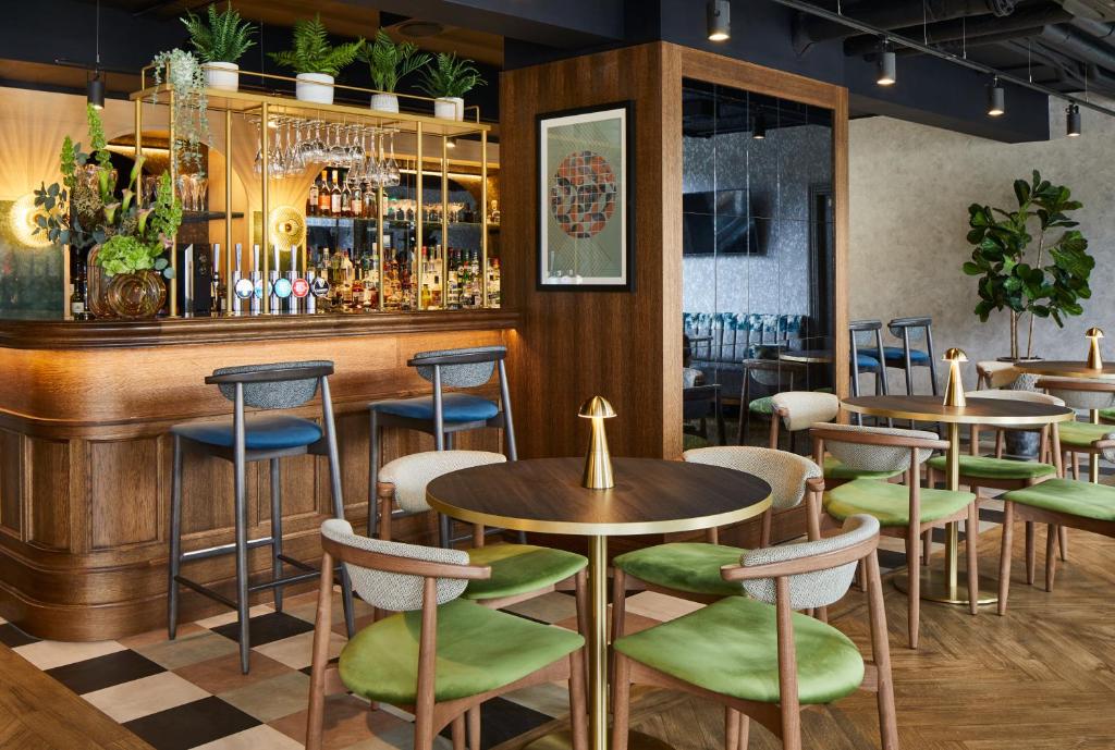 Leonardo Royal Hotel Edinburgh e um Bar moderno com mesas redondas, cadeiras verdes almofadadas, banquetas, copos pendurados e plantas em prateleiras. Ilustra o post de onde ficar em Edimburgo.