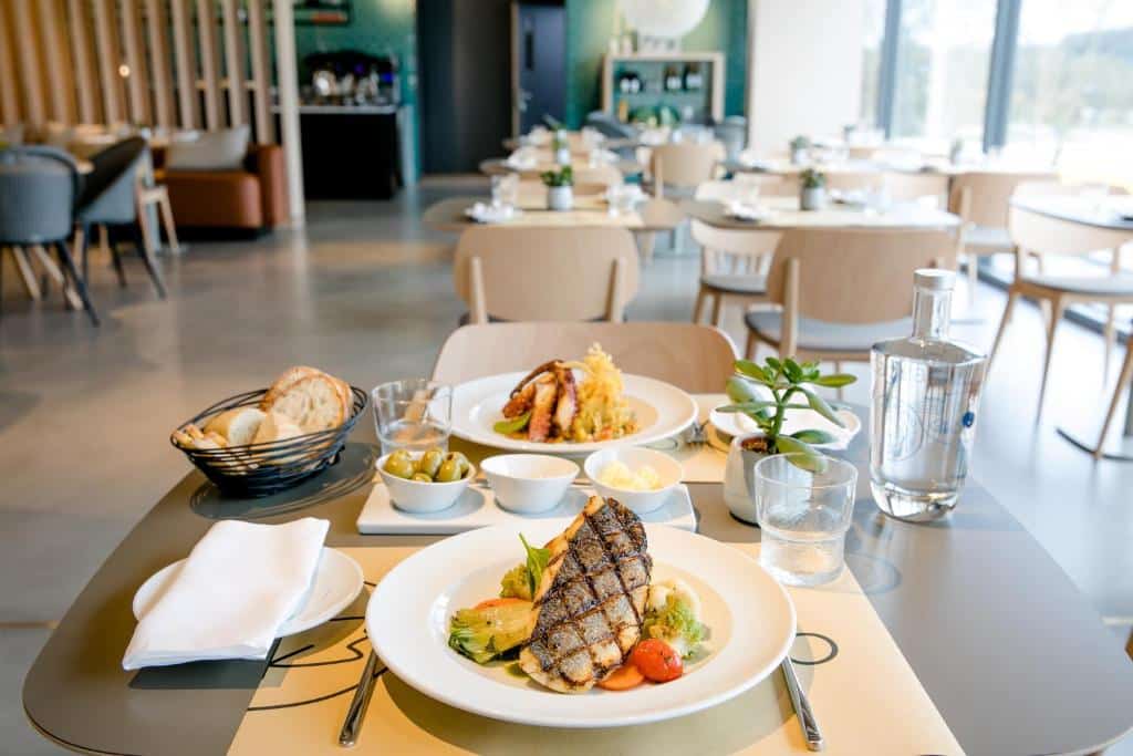 Uma mesa para duas pessoas com peixe grelhado, pão, azeitonas, sobremesa e água em uma área de jantar moderna e iluminada de um restaurante no Mercure Fátima.