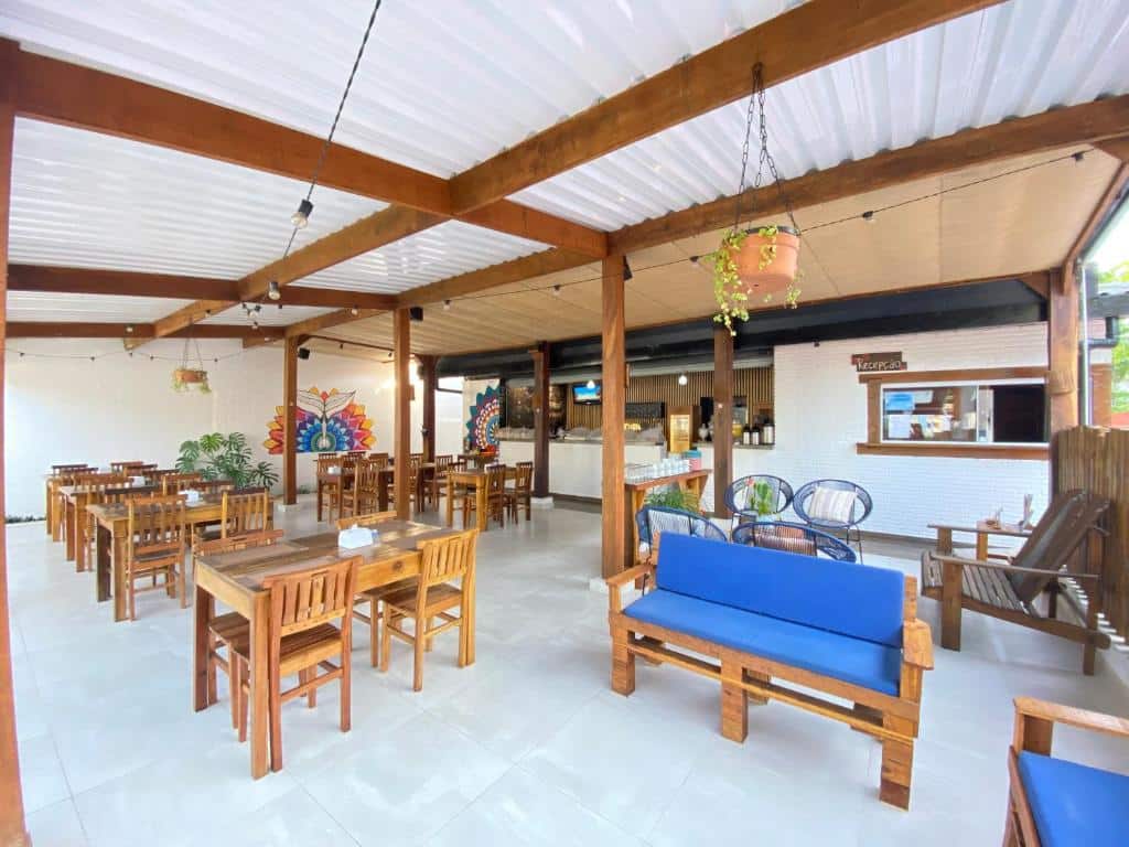 Restaurante ao ar livre com mesas de madeira, assentos almofadados azuis, piso de ladrilho branco e arte na parede ao fundo, na propriedade da Pousada Pono Village.
