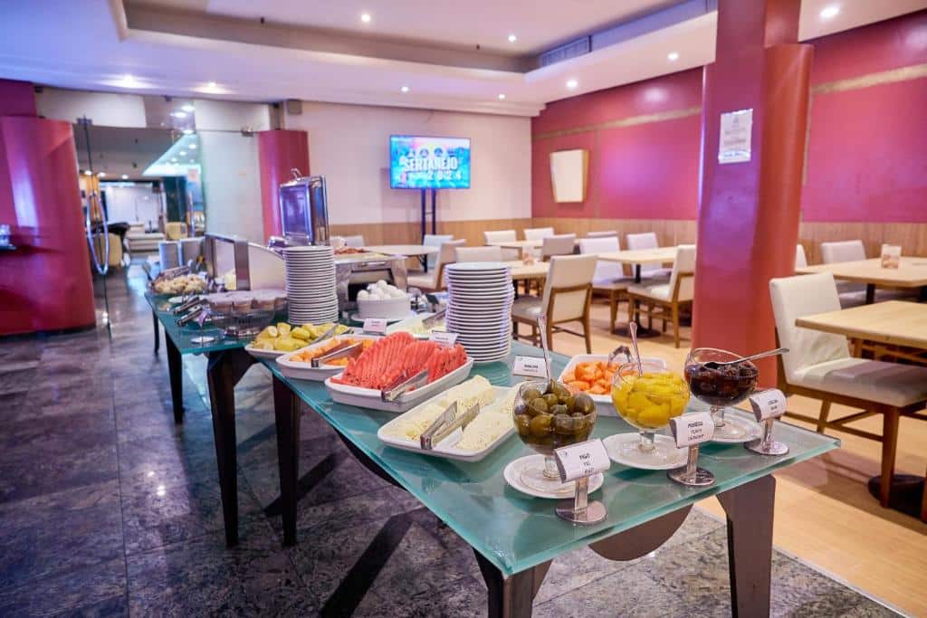 Uma mesa de bufê com pratos, frutas, azeitonas e outros itens alimentares em uma área de jantar espaçosa com mesas e cadeiras vazias, na propriedade do South American Copacabana Hotel.