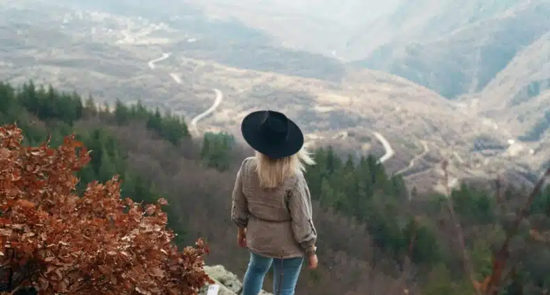 Uma pessoa de chapéu está em uma saliência rochosa, nas Rhodope Mountains, na Bulgaria, com vista para uma estrada sinuosa que atravessa um vale montanhoso arborizado. A foto é capa do post de eSIM Bulgária.
