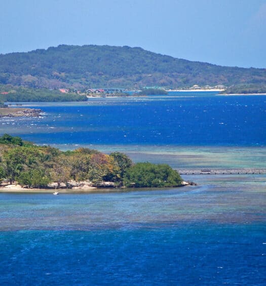Vista de Roatan com água azul, um píer, pequenas estruturas e uma colina verde coberta de árvores ao fundo. Imagem para ilustrar post de eSIM Honduras.