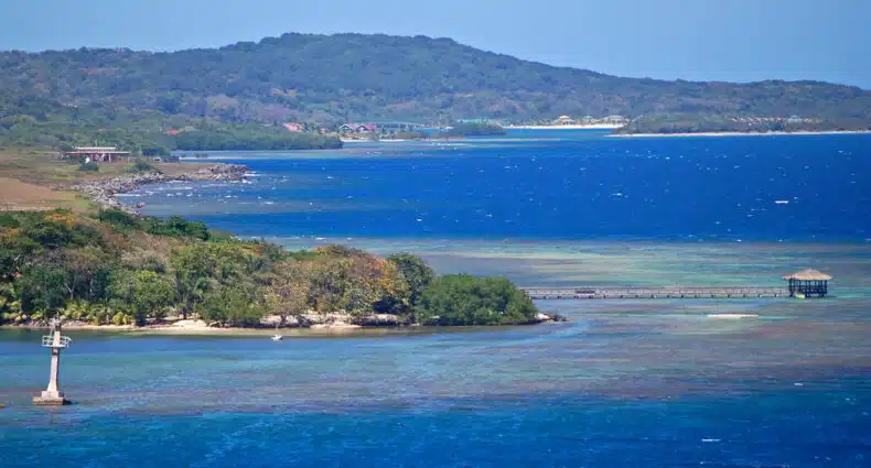 Vista de Roatan com água azul, um píer, pequenas estruturas e uma colina verde coberta de árvores ao fundo. Imagem para ilustrar post de eSIM Honduras.