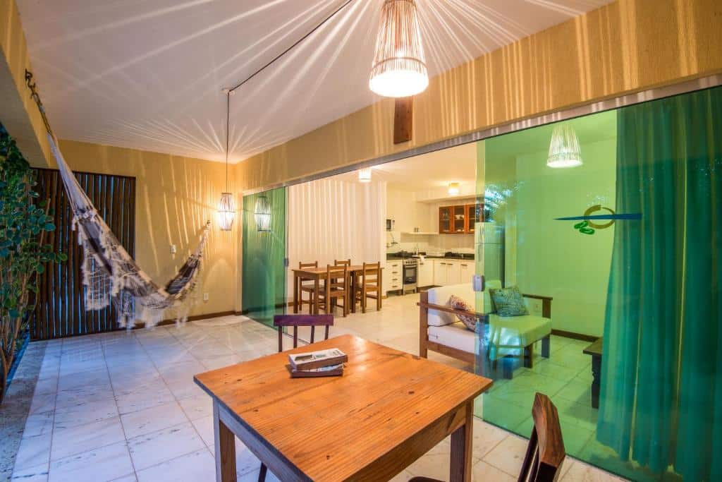Um pátio coberto com rede, mesa de madeira e cadeiras, que se abre para uma área de estar e cozinha com portas de vidro verde, no Apartamento no Village Imbassaí Breeze.