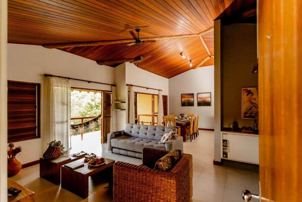 Uma sala de estar com teto de madeira, móveis de vime, um sofá cinza, janelas grandes e uma área de jantar ao fundo, no Apartamento dos Sonhos Imbassaí, para representar airbnb em Imbassaí.