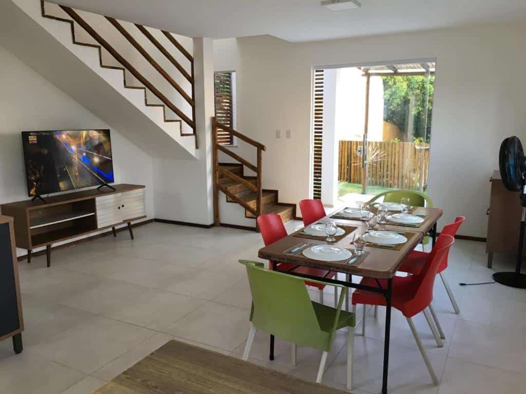 Uma área de jantar com uma mesa posta, cadeiras coloridas, uma TV ligada e uma escada que leva ao andar de cima em uma sala iluminada, na Casa duplex de temporada, para representar airbnb em Imbassaí.
