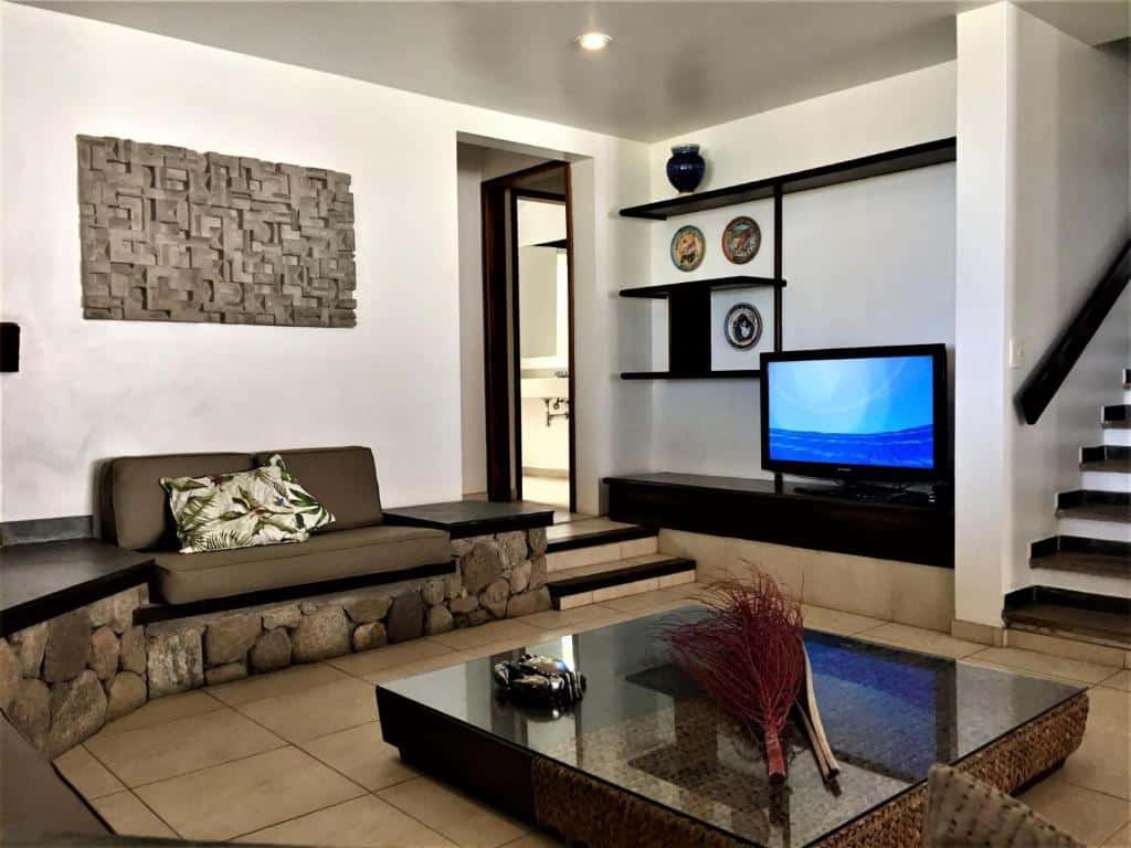 Sala de estar na Casa pé na areia de revista, com decoração de pedra e azulejo, uma TV, prateleiras na parede e um banco almofadado perto de uma escada, para representar airbnb na Praia da Baleia.