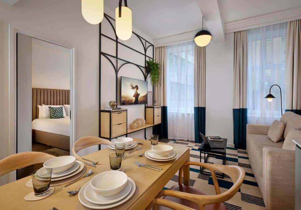 Uma moderna área de estar e jantar com mesa posta, sofá, TV e vista para um quarto com cama e travesseiro decorativo, no Citadines Holborn - Covent Garden London.