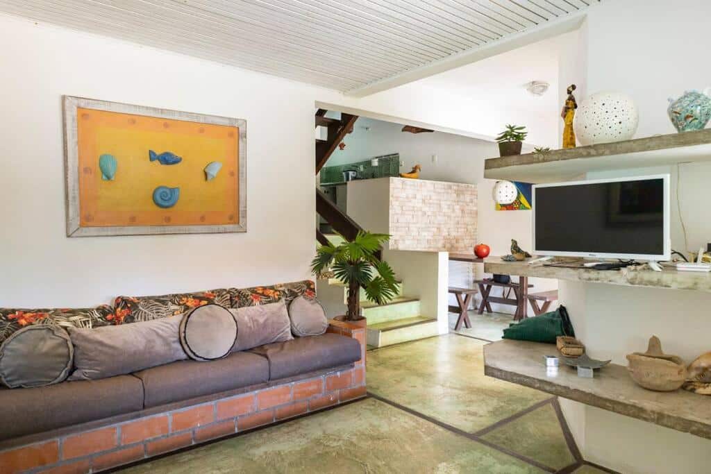 Uma sala de estar com um sofá cinza, arte na parede, uma TV em uma prateleira e uma vista da área de jantar e das escadas ao fundo, no Delba, para representar pousadas na Barra do Una.