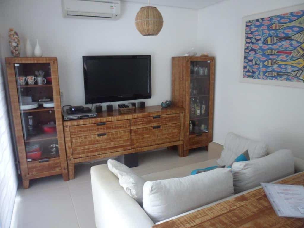 Sala de estar na Linda Casa em Juquehy com sofá branco, armários de madeira e suporte para TV, TV de parede, ar-condicionado e obras de arte com peixes coloridos, para representar airbnb na Praia da Baleia.