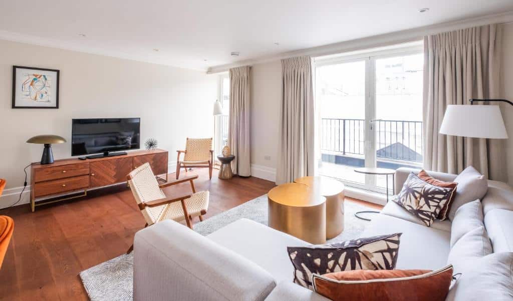 Sala de estar bem iluminada com um sofá seccional, TV, duas cadeiras, mesa de centro, janela grande e cortinas claras, no Native Mayfair.