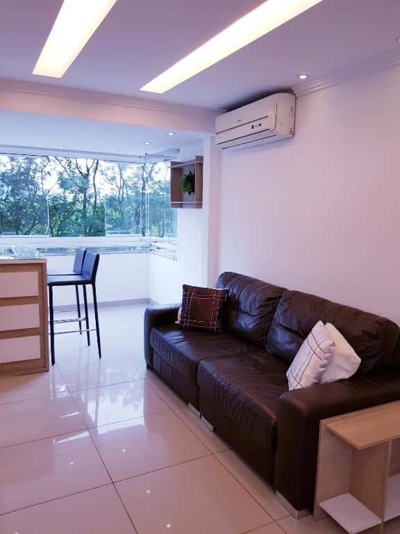 Sala de estar moderna com um sofá de couro marrom, mesa pequena, banquetas, janelas grandes e piso de ladrilho branco, no Rio Centro – Barra da Tijuca – Recreio – Praia.