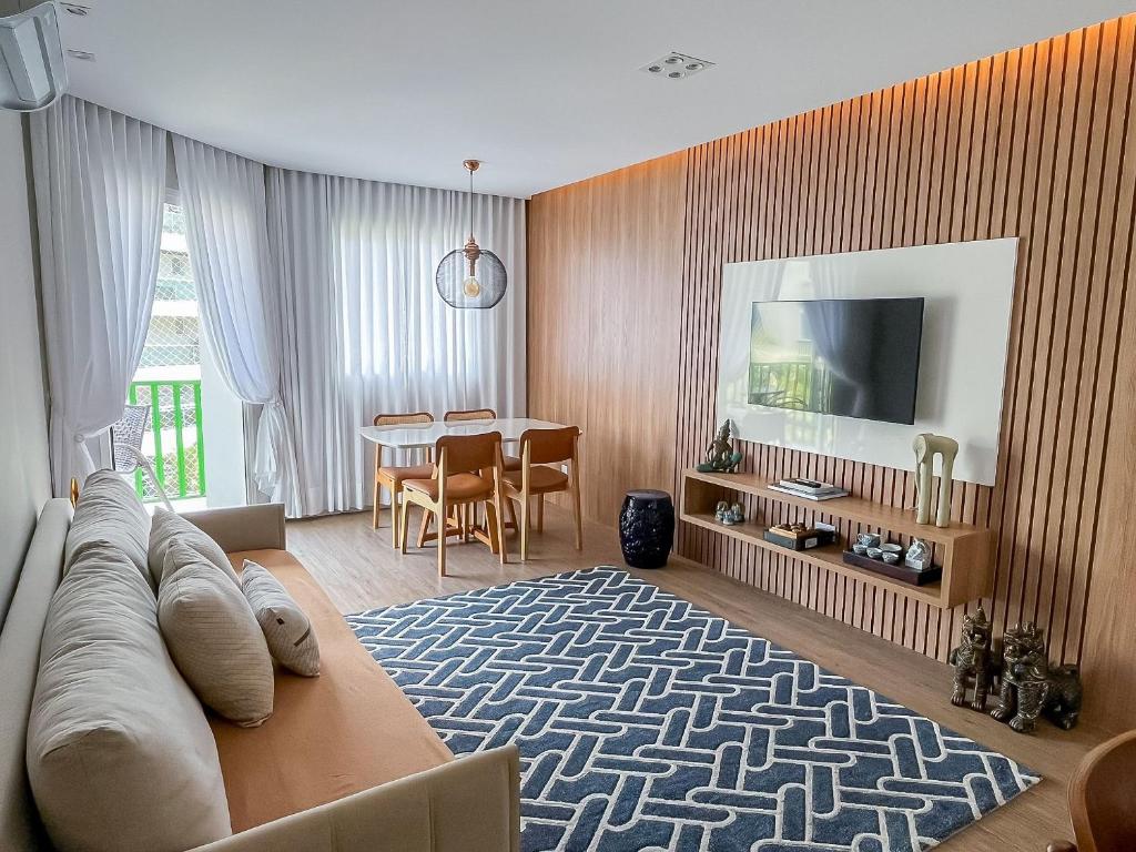 Sala de estar moderna com painéis de madeira, TV na parede, mesa de jantar, sofá bege e tapete azul geométrico, no apartamento do Sofisticado e Moderno - Enxoval Completo - Riviera.