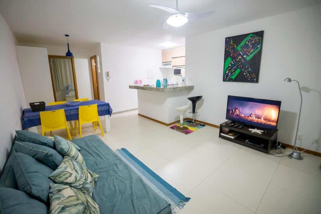 Uma sala de estar com um sofá azul-petróleo, TV, mesa de jantar com cadeiras amarelas, balcão de cozinha e decoração moderna, no Solaris Village.