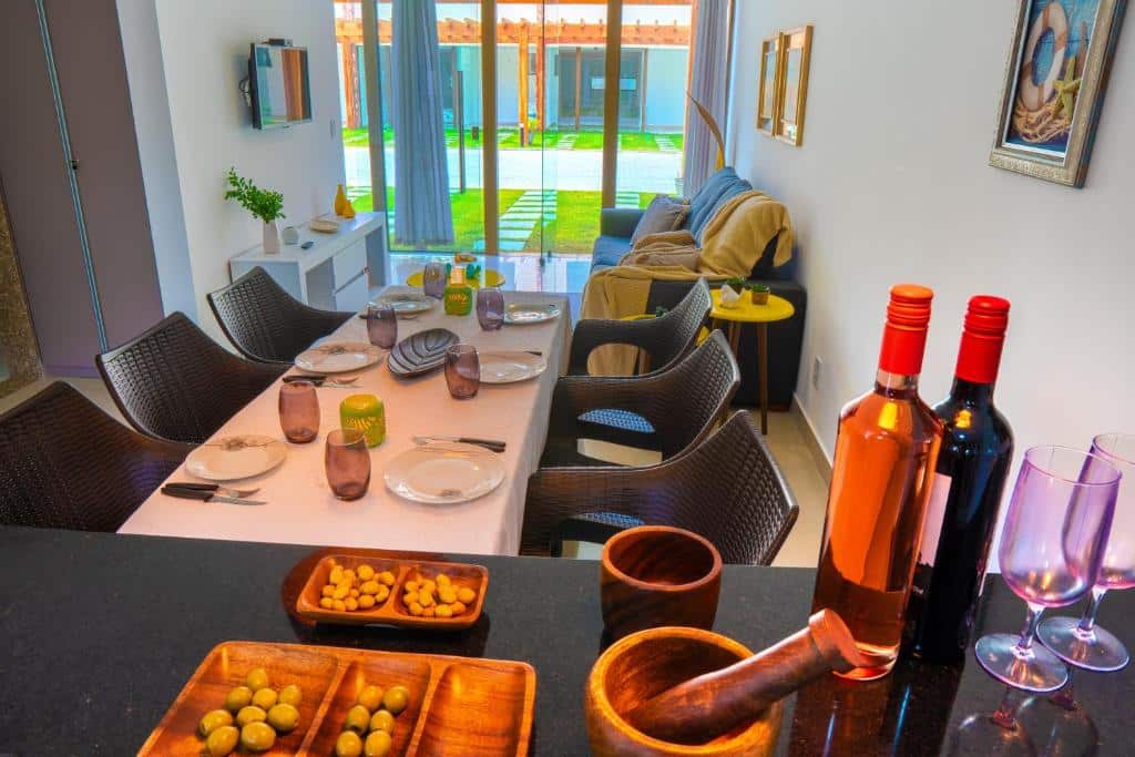 Uma mesa de jantar para seis pessoas com pratos, copos e talheres; garrafas de vinho e petiscos estão no balcão da cozinha em frente, no Village Imbassaí.