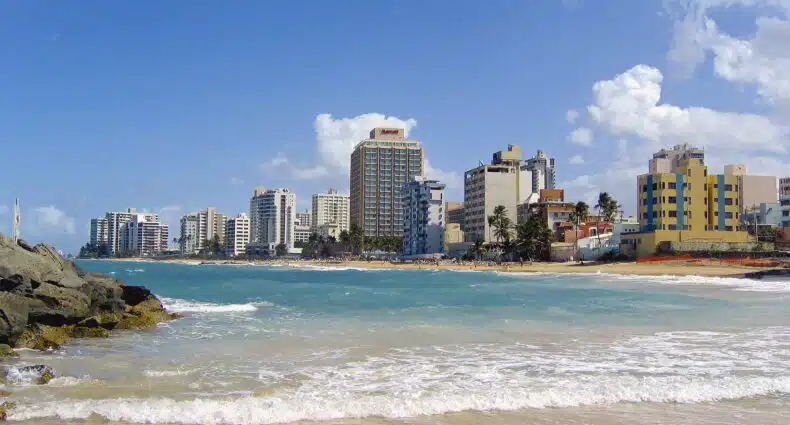 Praia de San Juan com prédios altos, praia arenosa, ondas do mar, pedras em primeiro plano e céu parcialmente nublado. Imagem para ilustrar post sobre eSIM Porto Rico.