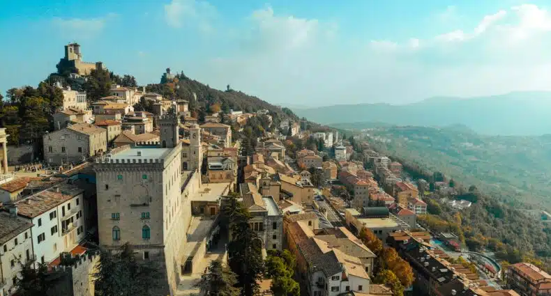 Vista aérea de uma cidade na encosta com edifícios de pedra e torres cercadas por colinas verdes sob um céu parcialmente nublado. Ilustra o eSIM San Marino.