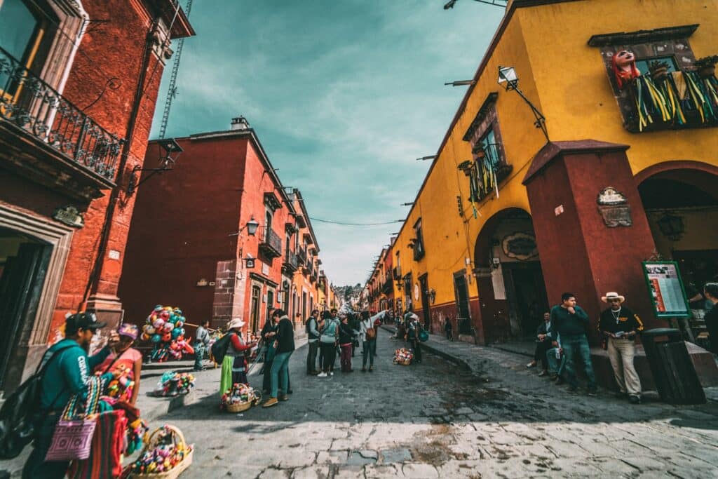 As pessoas caminham por uma rua de paralelepípedos ladeada por prédios coloridos vermelhos e amarelos e vendedores ambulantes. Ilustra o eSIM México.