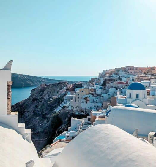 Edifícios brancos com cúpulas azuis têm vista para o mar em uma cidade costeira, com uma torre sineira em primeiro plano sob um céu claro que pode ser visitada com o eSIM Santorini.