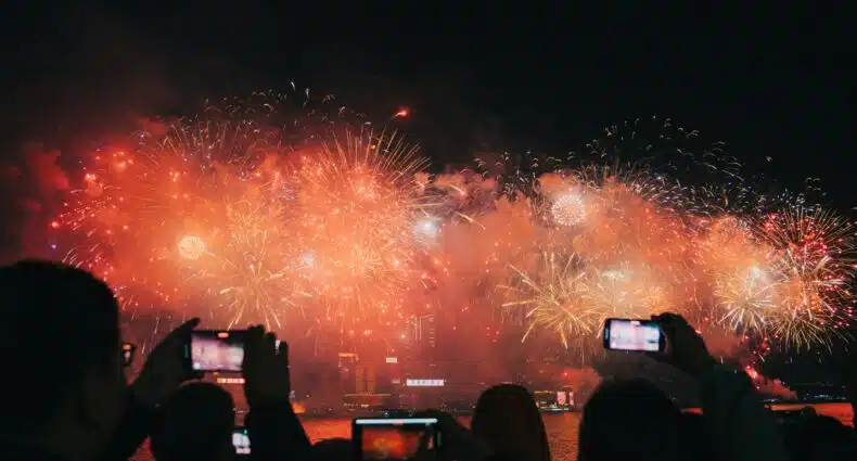 Pessoas assistem e gravam uma grande exibição de fogos de artifício sobre a água à noite, com muitas explosões iluminando o céu. É o Ano Novo Chinês ilustrando o eSIM Hong Kong