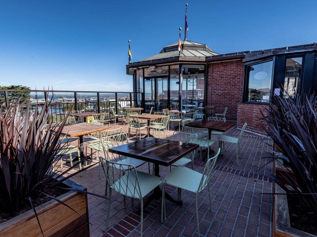 Pátio externo do restaurante com mesas de madeira, cadeiras de metal, piso de tijolos e vista para a cidade sob um céu azul claro, na propriedade do The Fairmont Heritage Place Ghirardelli Square.