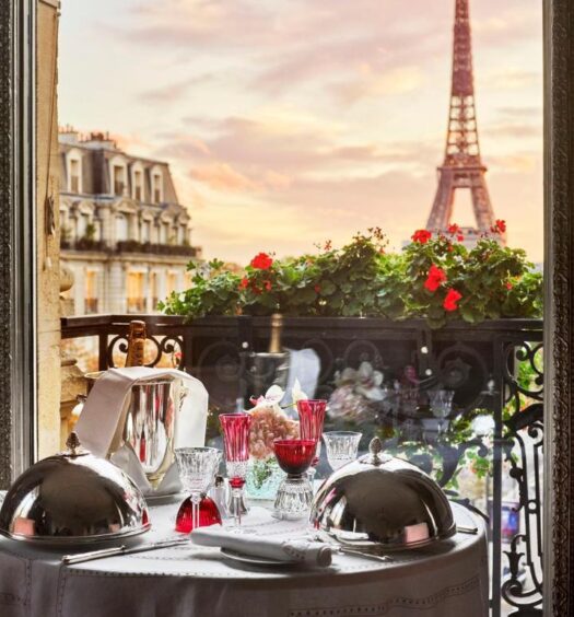 Uma mesa para dois com pratos cobertos e copos vermelhos tem vista para a Torre Eiffel ao pôr do sol por uma janela aberta, no Hôtel Plaza Athénée - Dorchester Collection, para representar hotéis românticos em Paris